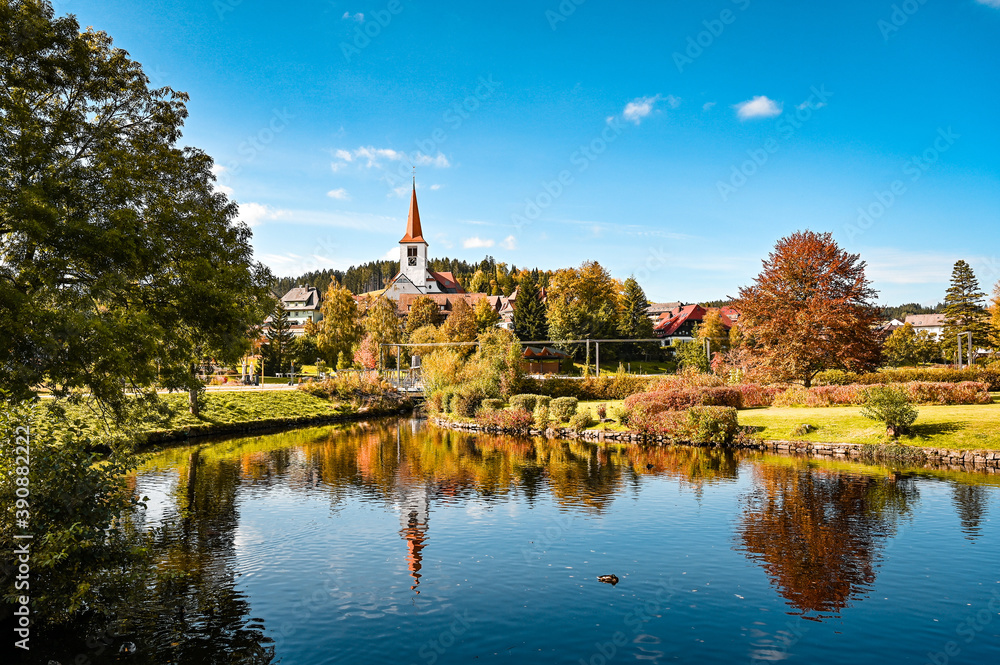 Cityscape view of the village of Schonach in the Black Forest of Germany during autumn. A church is in the centre and reflects in the water of a lake.