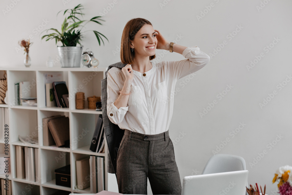 Successful female office worker in gray pants and light shirt with smile poses standing at workplace Successful female office worker in gray pants and light shirt with smile poses standing at workplace