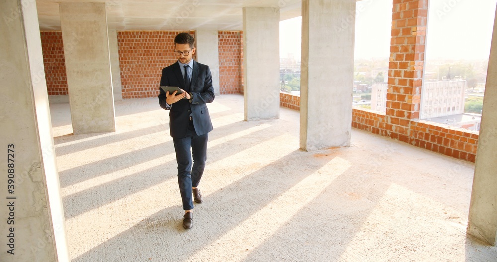 Caucasian male real-estate agent in glasses, suit and tie walking in building on construction phase, using tablet device and tapping or scrolling. Handsome businessman investor choosing mortgage.