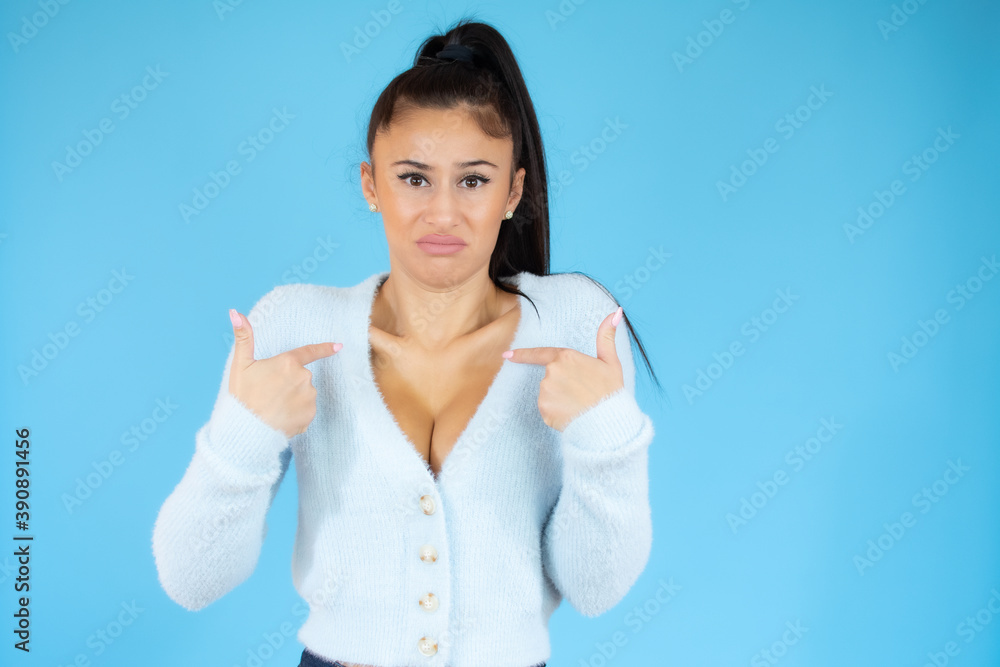 Portrait of young woman in casual blue sweater pointing fingers herself isolated over blue background Portrait of young woman in casual blue sweater pointing fingers herself isolated over blue background