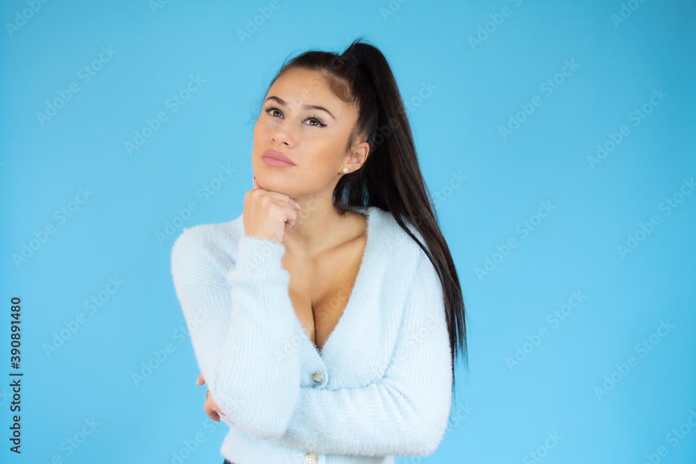 Young woman standing over blue isolated background with finger on face thinking about question, pensive expression. Doubt concept. Young woman standing over blue isolated background with finger on face thinking about question, pensive expression. Doubt concept.