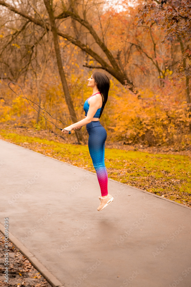 Young woman with jump rope in autumn park. Fitness female skipping before her workout outdoors. She wears bright sportswear.