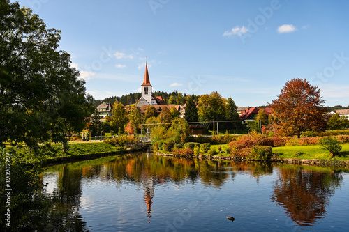Cityscape view of the village of Schonach in the Black Forest of Germany during autumn. A church is in the centre and reflects in the water of a lake.