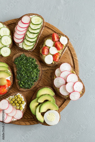 Vegetable sandwiches on the wood board on the gray background. Fresh bread with pesto, cucumber, avocado, cherry tomatoes, radish and cheese. Delicious breakfast. Flat lay, top view.