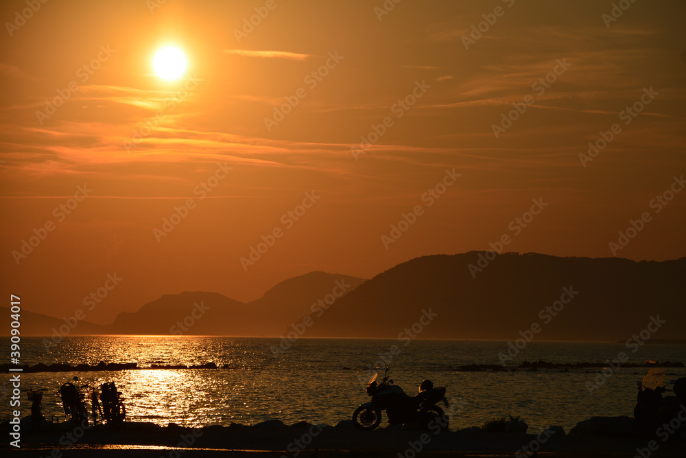 Sunset on the beach. The mountains in the background.  In the foreground, silhouettes of motorcycles. 