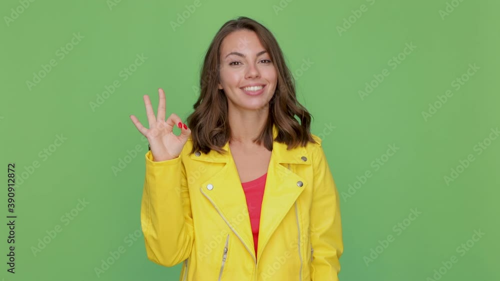Cheerful funny young brunette woman 20s years old in casual yellow leather jacket posing isolated on green background in studio. People lifestyle concept. Looking camera showing Ok gesture blinking Cheerful funny young brunette woman 20s years old in casual yellow leather jacket posing isolated on green background in studio. People lifestyle concept. Looking camera showing Ok gesture blinking