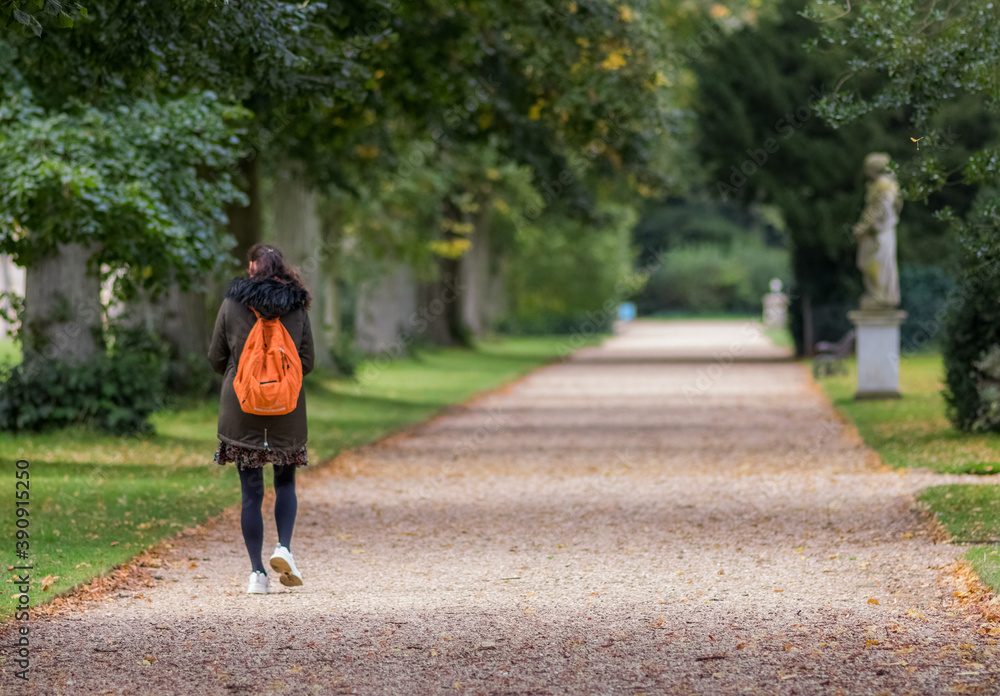 woman walking away from camera along gravel path with orange rucksack, the distance is blurred woman walking away from camera along gravel path with orange rucksack, the distance is blurred