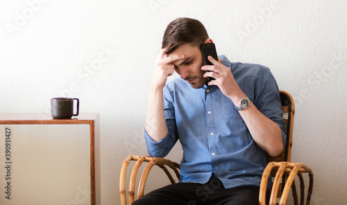Retrato de un hombre adulto caucásico y apuesto sentado sobre un sillón de mimbre dentro de un cálido apartamento preocupado mientras habla desde su teléfono móvil.
