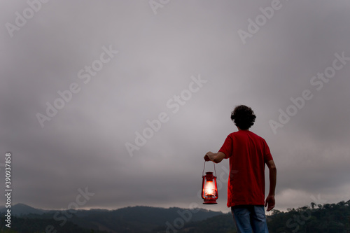 Young man in red t-shirt, holding a kerosene lamp to light his way, with mountains in the background.