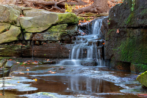 The blurred water of Little Davis Falls in Hamilton, Ontario cascades and reflects into a pool below, dotted with colourful fallen Autumn leaves.