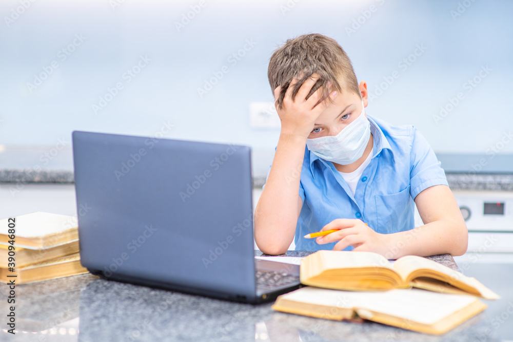 A boy studies remotely near a laptop in a medical mask, leaning over open books at home in the kitchen, wearily put his head on his palm. Education during the coronavirus pandemic