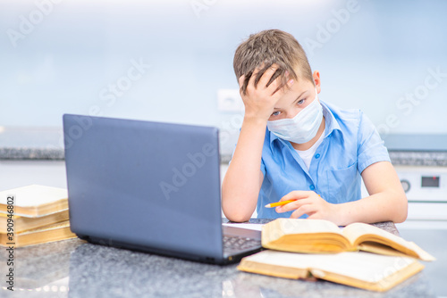 A boy studies remotely near a laptop in a medical mask, leaning over open books at home in the kitchen, wearily put his head on his palm. Education during the coronavirus pandemic