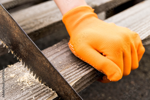 A hand in an orange glove is sawing a board with an old rusty hand saw.