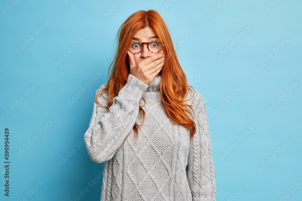 Young shocked redhead young woman covers mouth and stares stupefied hears  embarrassing news wears transparent glasses grey knitted sweater isolated  over blue background. Hiding secret concept Stock Photo