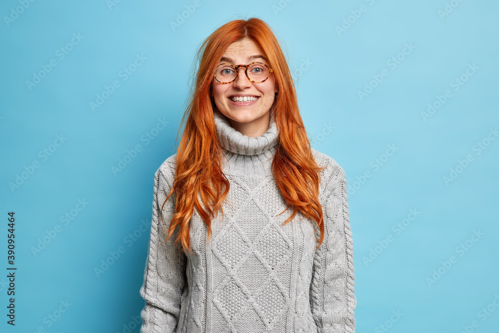 Lovely redhead young woman with european appearance smiles pleasantly dressed in warm sweater expresses positive emotions hears good news isolated over blue background. Happy emotions concept Lovely redhead young woman with european appearance smiles pleasantly dressed in warm sweater expresses positive emotions hears good news isolated over blue background. Happy emotions concept