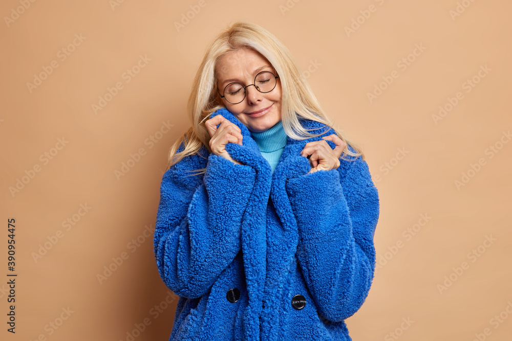 Photo of fashionable blonde woman tilts head and closes eyes wears round spectacles blue fur coat recalls something pleasant isolated over brown background. Winter fashion and style concept. Photo of fashionable blonde woman tilts head and closes eyes wears round spectacles blue fur coat recalls something pleasant isolated over brown background. Winter fashion and style concept.