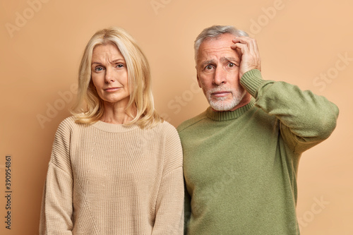 Фотография Tired displeased blonde wrinkled woman looks calmly at camera and embarrassed troublesome man looks with panic and fearful expression isolated over brown wall