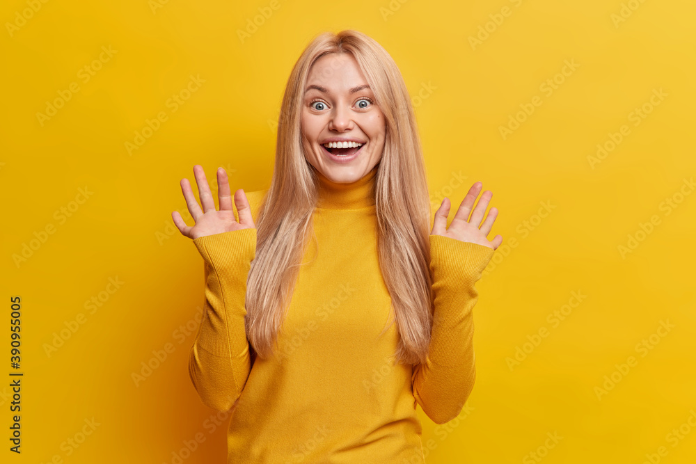 Charming blonde Caucasian woman feels very glad raises hands and smiles broadly notices something awesome or thrilling dressed in casual clothes isolated over yellow background. Happy emotions Charming blonde Caucasian woman feels very glad raises hands and smiles broadly notices something awesome or thrilling dressed in casual clothes isolated over yellow background. Happy emotions