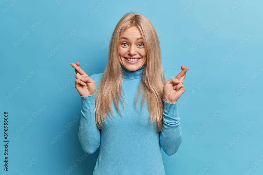 Cheerful smiling blonde woman waits for special moment keeps fingers crossed wishes good luck dressed in casual turtleneck isolated over blue background aniticipates something with great desire Cheerful smiling blonde woman waits for special moment keeps fingers crossed wishes good luck dressed in casual turtleneck isolated over blue background aniticipates something with great desire