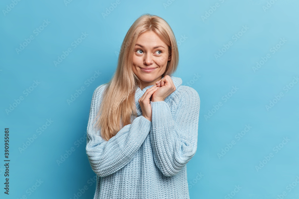 Happy dreamy blonde young woman keeps hands together and concentrated away with pleasant smile contemplates something beautiful enjoys view wears warm knitted jumper isolated over blue wall. Happy dreamy blonde young woman keeps hands together and concentrated away with pleasant smile contemplates something beautiful enjoys view wears warm knitted jumper isolated over blue wall.
