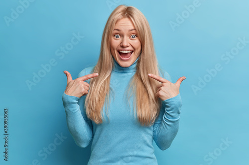 Confident cheerful blonde woman looks amazed overemotive expression points at herself suggests own help says pick me dressed in casual turtleneck isolated over blue background. This is my personality