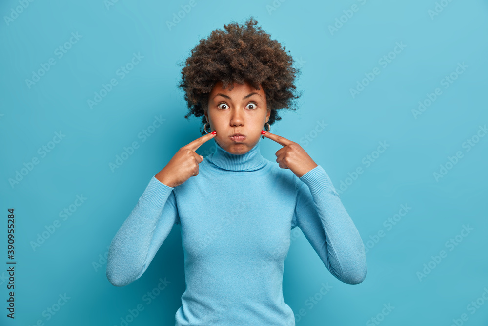 Studio shot of funny dark skinned woman blows air and points index fingers at cheeks makes funny grimace holds breath dressed in casual turtleneck isolated over blue background foolishes around Studio shot of funny dark skinned woman blows air and points index fingers at cheeks makes funny grimace holds breath dressed in casual turtleneck isolated over blue background foolishes around