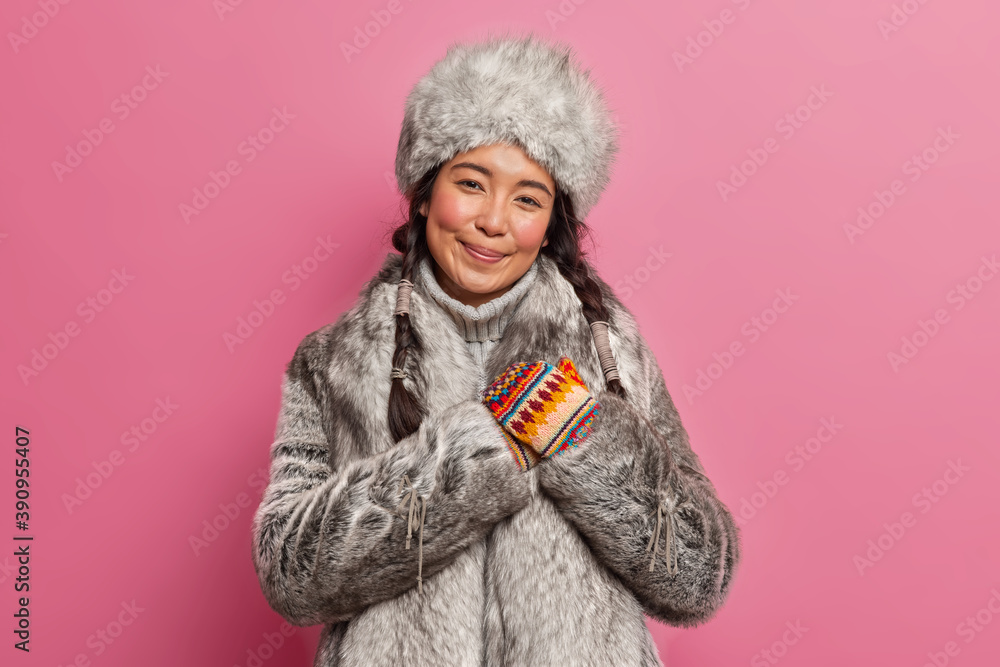 Pleased brunette woman inhabitant of far north makes gratitude gesture smiles gently wears grey fur winter outfit poses against rosy background. Touched arctic lady from far north being thankful Pleased brunette woman inhabitant of far north makes gratitude gesture smiles gently wears grey fur winter outfit poses against rosy background. Touched arctic lady from far north being thankful