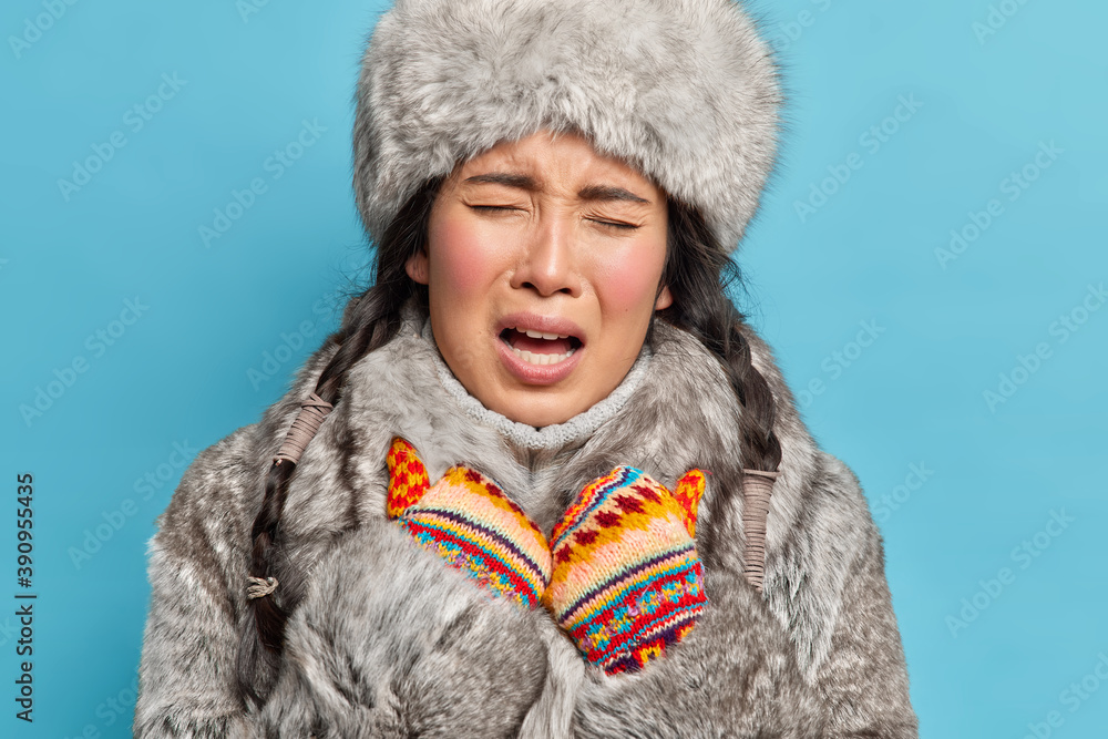Close up shot of displeased young Asian inuit woman wears knitted mittens  and winter outfit feels cold during frosty winter isolated over blue  background. Arctic female inhabitant of far north Photos