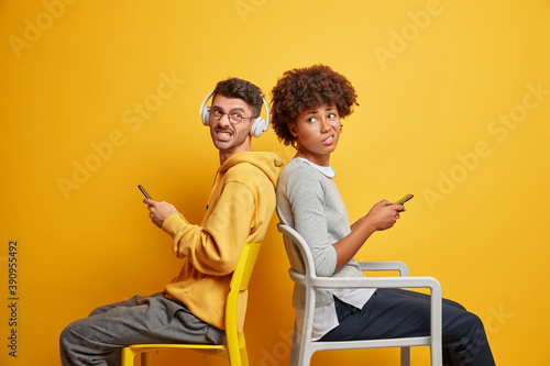 Two mixed race friends wait for something sit back to each other on chairs smirk faces and have irritated expressions dressed in casual clothes isolated over yellow background. Technology users © wayhome.studio 