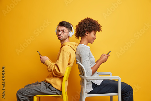 Woman and man sit back to each other on chairs dont speak use modern cellulars communicate online isolated over yellow background. Intterracial friends addicted to technologies and internet. photo