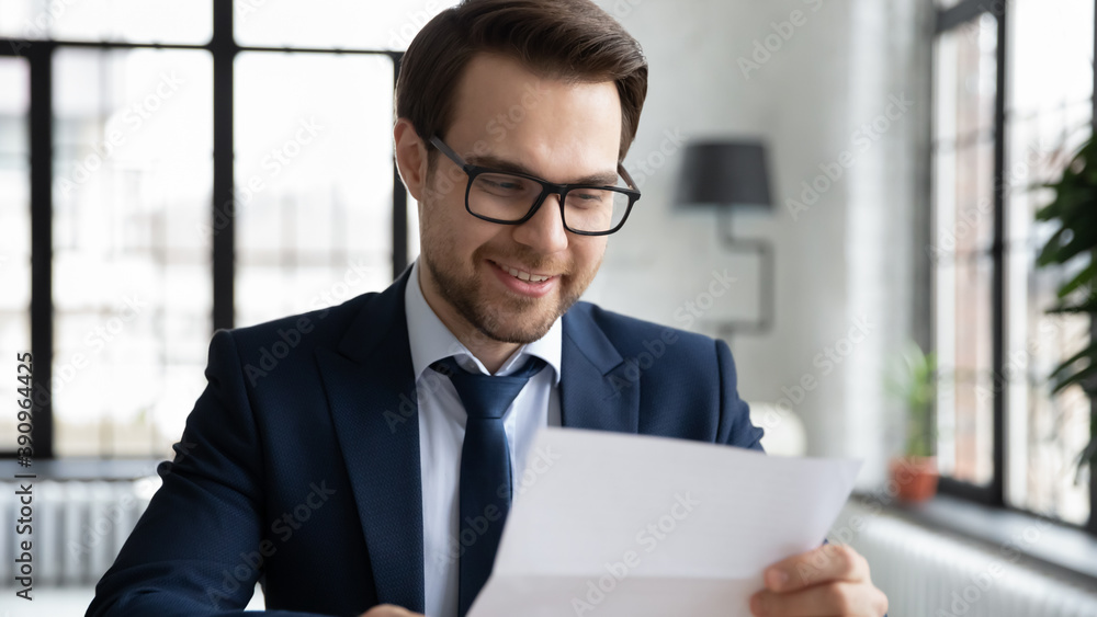 Head shot smiling young executive manager in formal wear and glasses reading pleasant news in correspondence, satisfied with project results financial report, successful worker consider paper document Head shot smiling young executive manager in formal wear and glasses reading pleasant news in correspondence, satisfied with project results financial report, successful worker consider paper document