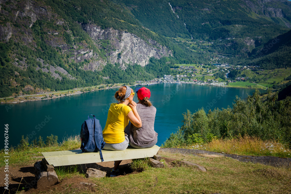 Fototapeta premium mom and daughter sit on the bench with their backs. Norway, view from the mountain to the city of Geiranger and the fjord. Mom and daughter together.