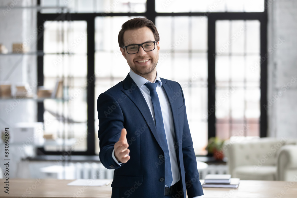 Happy young businessman in glasses looking at camera, reaching out hand making welcoming gesture or ready handshaking with partner or colleague, smiling banker proposing good deal to client. Happy young businessman in glasses looking at camera, reaching out hand making welcoming gesture or ready handshaking with partner or colleague, smiling banker proposing good deal to client.