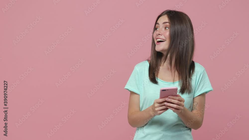 Surprised young woman 20s years old in blue t-shirt posing isolated on pink background studio. People lifestyle concept. Using mobile cell phone typing sms message say wow point aside showing thumb up Surprised young woman 20s years old in blue t-shirt posing isolated on pink background studio. People lifestyle concept. Using mobile cell phone typing sms message say wow point aside showing thumb up