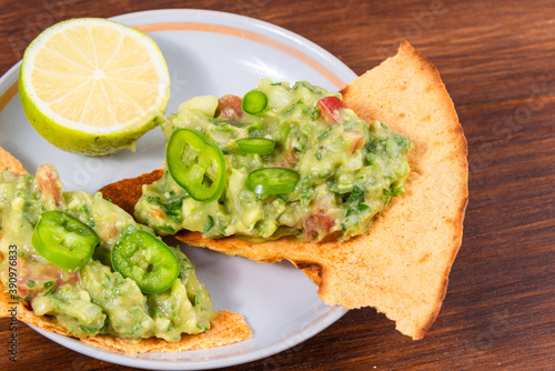 Homemade guacamole - a traditional Mexican appetizer of avacado, onion and tomato on toasted tortilla pieces in a plate with half a lime on a rustic wooden table