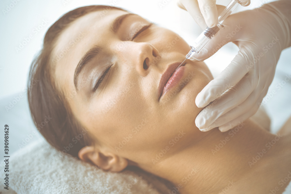 Naklejka premium Beautician doing beauty procedure with syringe to face of young brunette woman in sunny clinic. Cosmetic medicine and surgery, beauty injections
