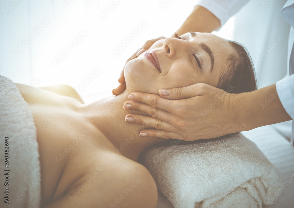 Beautiful brunette woman enjoying facial massage with closed eyes in sunny spa center. Relaxing treatment and cosmetic medicine concepts