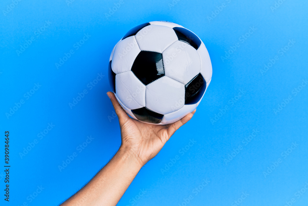 Hand of young hispanic man holding soccer ball over isolated blue background.