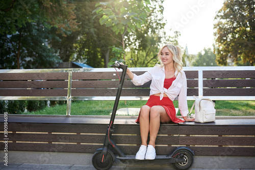 Young blond woman, sitting on bench with electric scooter in city center. Female, wearing red skirt and white top,posing for portrait Summer leisure activity. Traveler exploring the town on scooter.