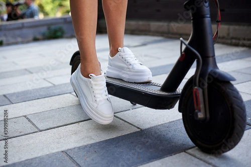 Close-up picture of woman's legs, riding on electric scooter in city center. Female, wearing white sneakers. Summer leisure activity. Traveler exploring the town, spending time outdoors.