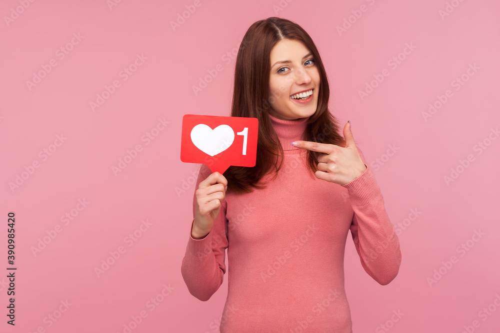 Smiling brunette woman in pink sweater pointing finger at sign with like counter, blogger waiting for feedback and positive rate. Indoor studio shot isolated on pink background Smiling brunette woman in pink sweater pointing finger at sign with like counter, blogger waiting for feedback and positive rate. Indoor studio shot isolated on pink background