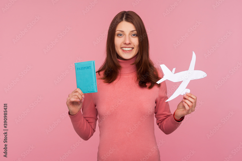 Happy brunette woman in pink sweater holding paper plane model and passport in hands looking at camera with toothy smile. Indoor studio shot isolated on pink background Happy brunette woman in pink sweater holding paper plane model and passport in hands looking at camera with toothy smile. Indoor studio shot isolated on pink background
