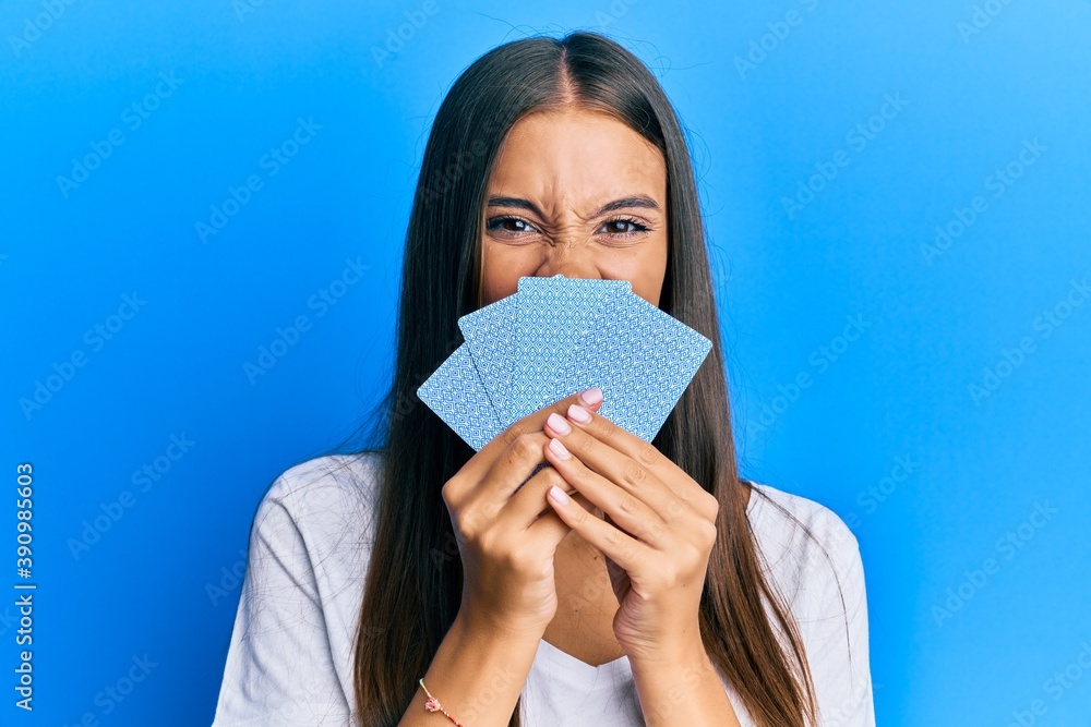 Young hispanic woman playing gambling poker covering face with cards sticking tongue out happy with funny expression. Young hispanic woman playing gambling poker covering face with cards sticking tongue out happy with funny expression.