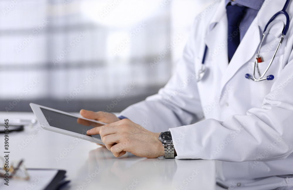 Unknown male doctor sitting and working with tablet computer in clinic at his working place, close-up. Young physician at work. Perfect medical service, medicine concept Unknown male doctor sitting and working with tablet computer in clinic at his working place, close-up. Young physician at work. Perfect medical service, medicine concept