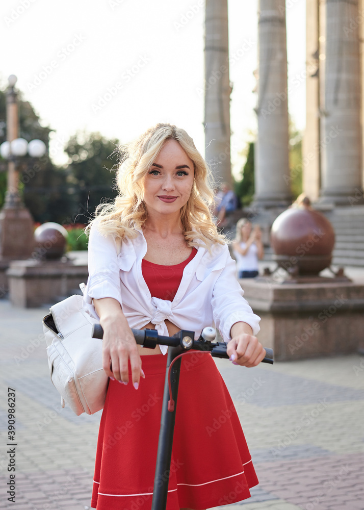 Young blond woman, posing with black electric scooter in city center in front of old historical building with pillars. Summer leisure activity. Traveler exploring the town, spending time outdoors.