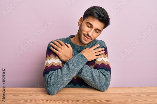 Young handsome man wearing casual winter sweater sitting on the table hugging oneself happy and positive, smiling confident. self love and self care © Krakenimages.com