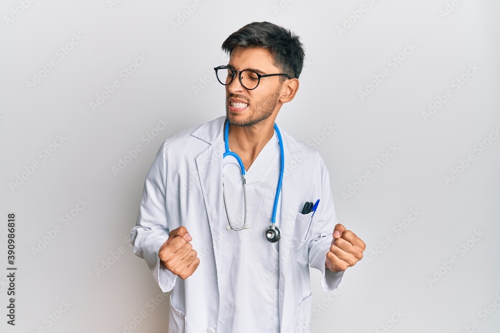 Young handsome man wearing doctor uniform and stethoscope excited for success with arms raised and eyes closed celebrating victory smiling. winner concept. Young handsome man wearing doctor uniform and stethoscope excited for success with arms raised and eyes closed celebrating victory smiling. winner concept.