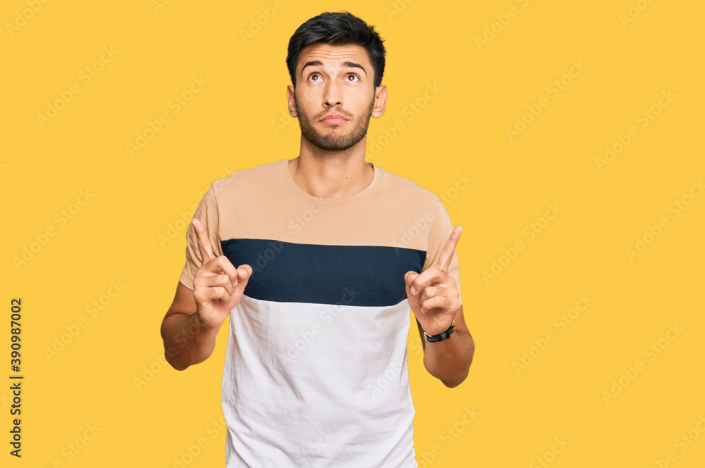 Young handsome man wearing casual clothes pointing up looking sad and upset, indicating direction with fingers, unhappy and depressed.