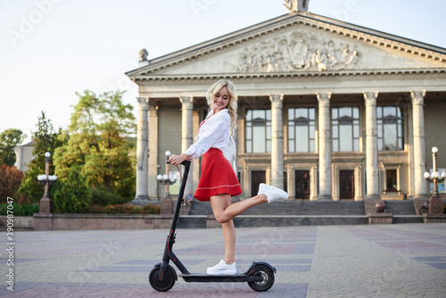 Young blond woman, riding on black electric scooter in city center in front of old historical building with pillars. Summer leisure activity. Spending free time outdoors when weather is warm.
