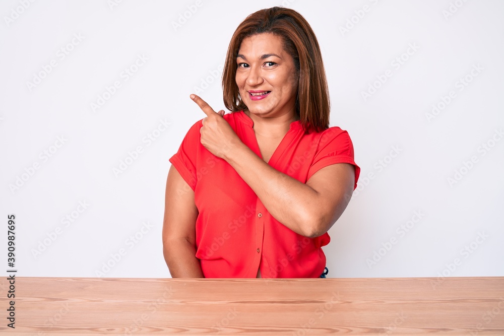 Middle age brunette hispanic woman wearing casual clothes sitting on the table smiling cheerful pointing with hand and finger up to the side Middle age brunette hispanic woman wearing casual clothes sitting on the table smiling cheerful pointing with hand and finger up to the side